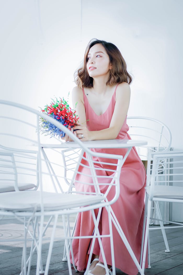 A woman in a pink dress holding colorful flowers while sitting indoors on white chairs.